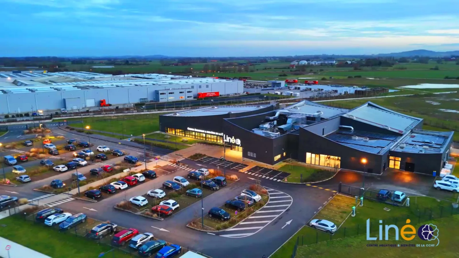 Centre aquatique de Wormhout (Linéo) vu par drone à la tombée du jour : bâtiment éclairé, parking et zone d'activités en arrière-plan